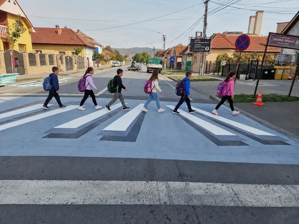 Mai multe persoane cu rucsacuri traversează o trecere pietonală cu benzi albe și forme gri, între clădiri și indicatoare rutiere.