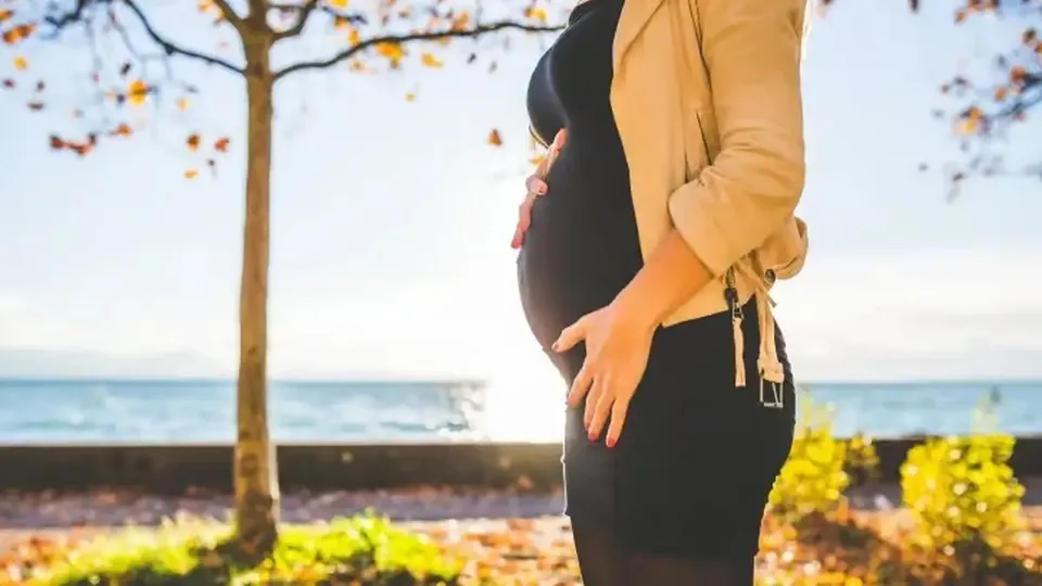 O persoană cu abdomen proeminent poartă o rochie neagră și un cardigan bej, cu mâinile așezate pe abdomen; în fundal se văd un copac și apă.