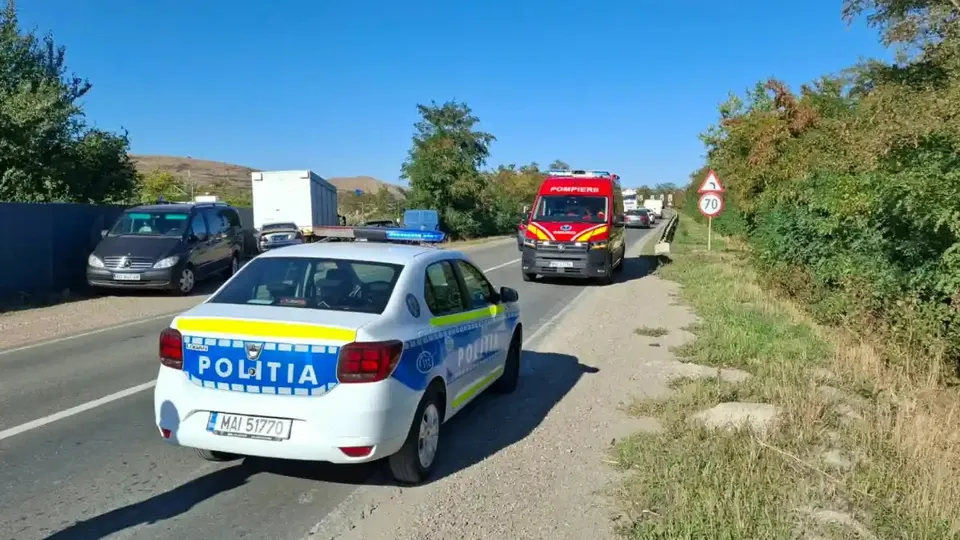 O mașină albă de poliție și o ambulanță roșie se află pe un drum asfaltat, lângă alte vehicule, vegetație și indicatoare rutiere.
