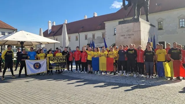 Un grup de persoane în haine sportive colorate stă pe o piață pavată, ținând steaguri și bannere în fața unui monument.