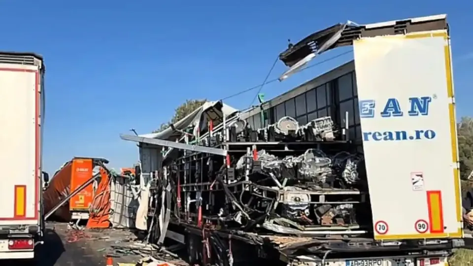 Un camion cu remorca albă este grav avariat, cu structura laterală expusă; în stânga se văd un camion portocaliu și partea din spate a unei alte remorci.