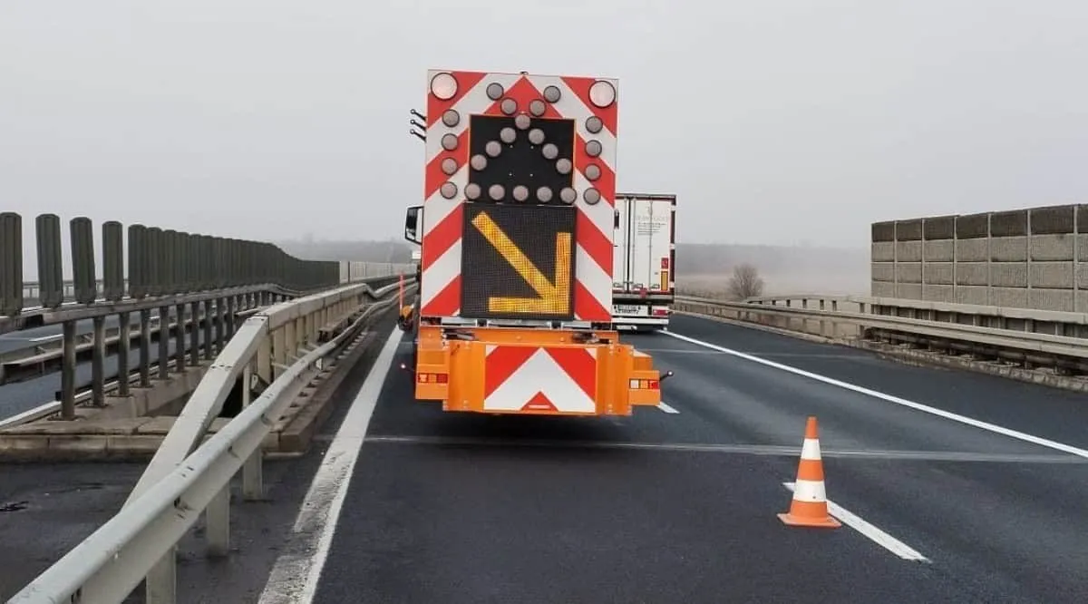 Un vehicul cu panou luminos și marcaje roșii și albe se află pe carosabil, lângă un con portocaliu și parapete metalice.