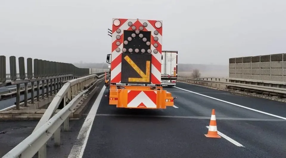 Un vehicul cu panou luminos și marcaje roșii și albe se află pe carosabil, lângă un con portocaliu și parapete metalice.