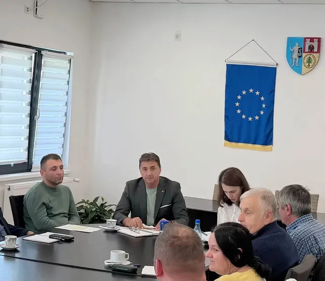 Mai multe persoane sunt așezate în jurul unei mese cu documente, pahare și dispozitive, într-o încăpere cu steagul Uniunii Europene pe perete.