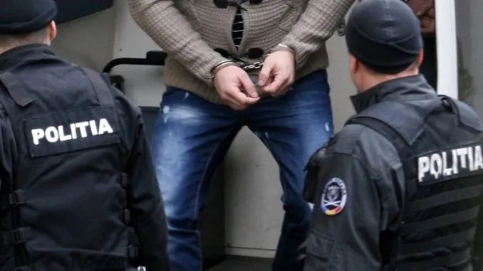 Două persoane în uniforme închise la culoare, cu inscripția „POLIȚIA”, stau lângă o persoană în blugi și pulover, cu mâinile împreunate.